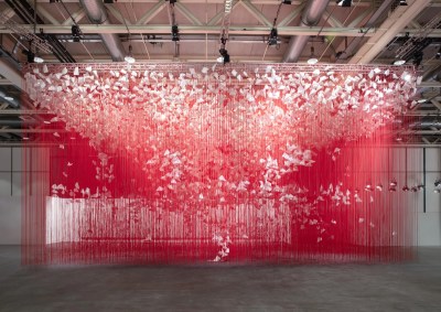 An installation composed of hanging red strings strewn with sheets of paper that appear to fly among them.