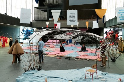 A group of people milling around a ovular structure whose floor is covered in pink and blue mats. A person nearby speaks into a megaphone.