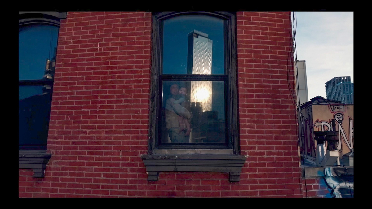 A film still showing a mother holding her child. She is seen from a window in a red-brick building. In the window's reflection is a high-rise gleaming in the sun.