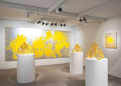 Sculptures composed of golden beads that are set atop white plinths. They are shown before paintings of yellow blotches against a grey background.