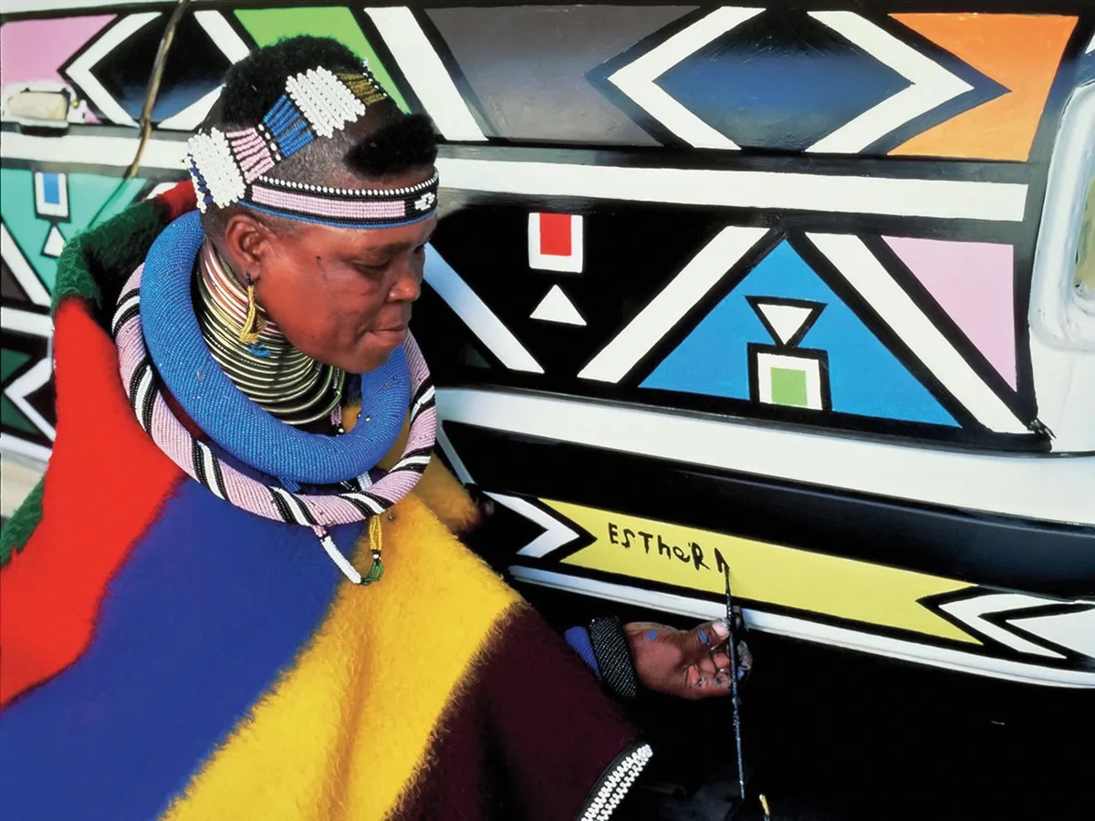 A Ndebele woman in raditional bearded garb is painting her name, Esther, on a geometric abstraction.