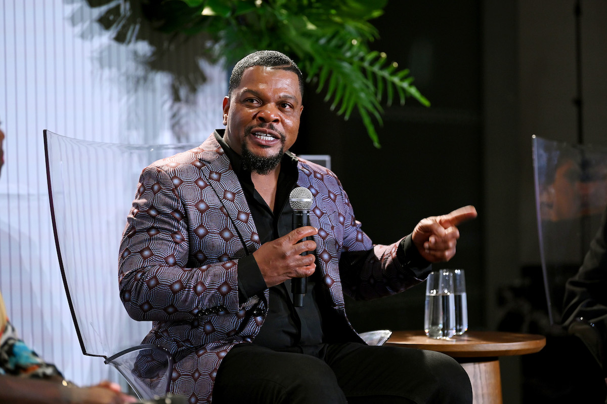 A Black man speaking into a microphone beside glasses of water. He points one finger. He wears a purple jacket with diamond-shaped patterning on it.