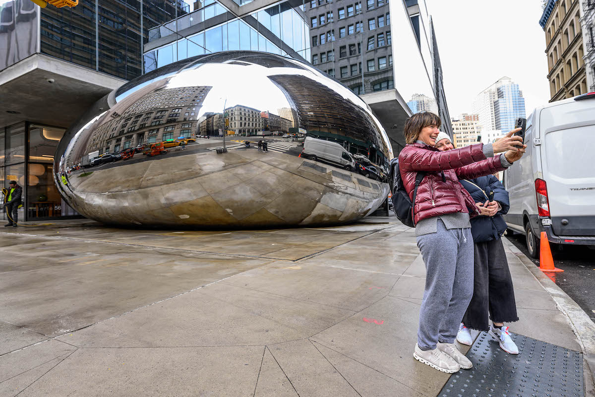 Two women take a selfie with a sculpture resembling a giant metallic bean being squashed beneath a luxury building.