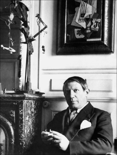 Pablo Picasso c. 1930, with his painting Still Life: Guitar, Newspaper, Glass, and Ace of Clubs (1914) in the background.