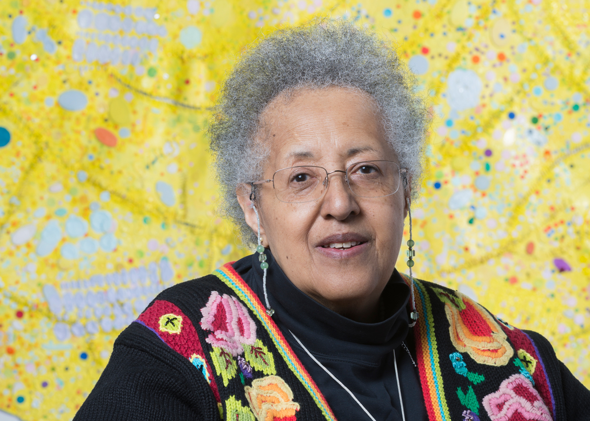 Portrait of Howardena Pindell, an Older Black woman who sits in front of an abstract painting that is mostly yellow.