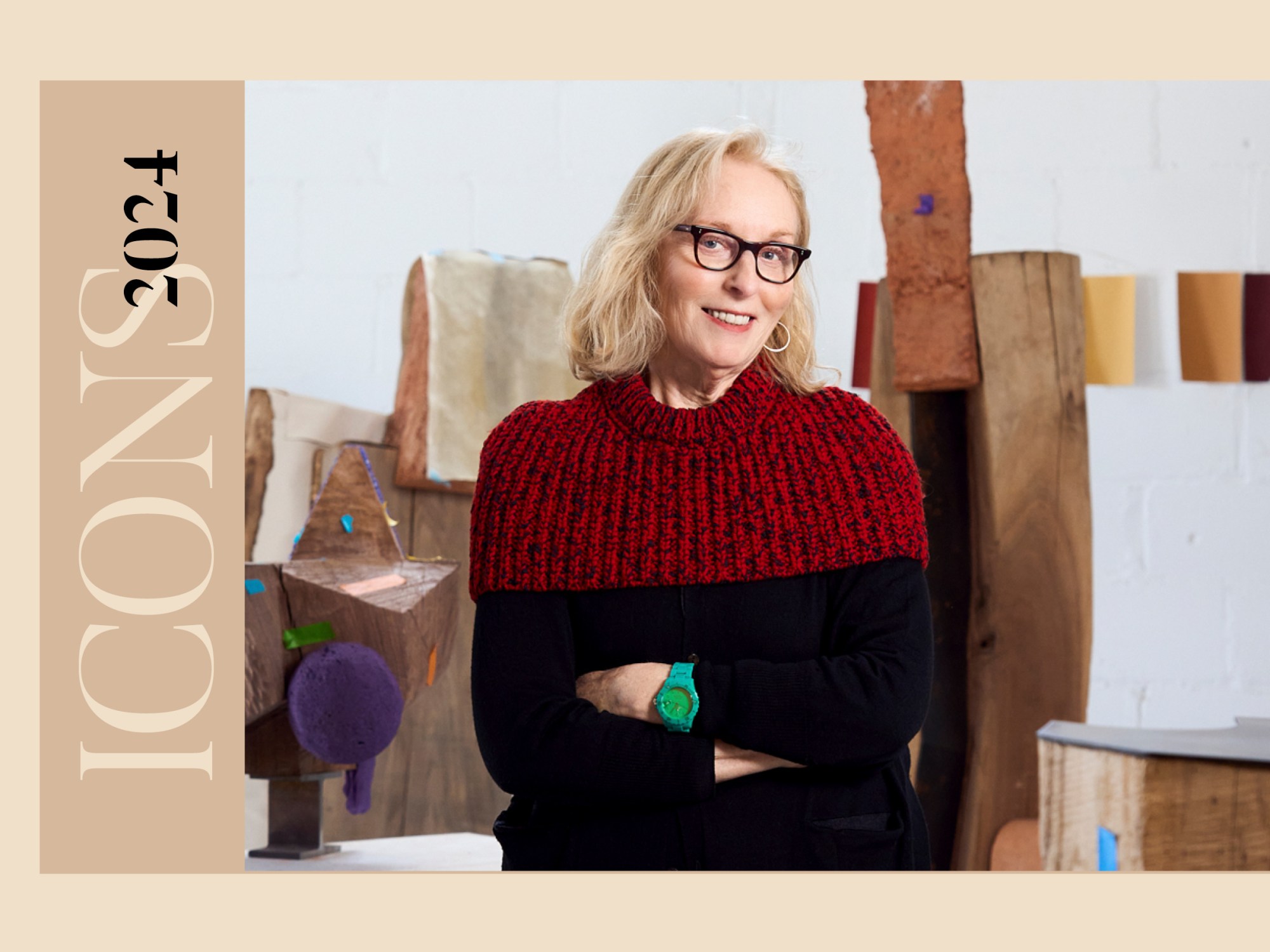 An older while woman with shoulder-length blonde hair, wearing a black and red sweater, has her arms crossed and is standing in a space with wood sculptures in the background