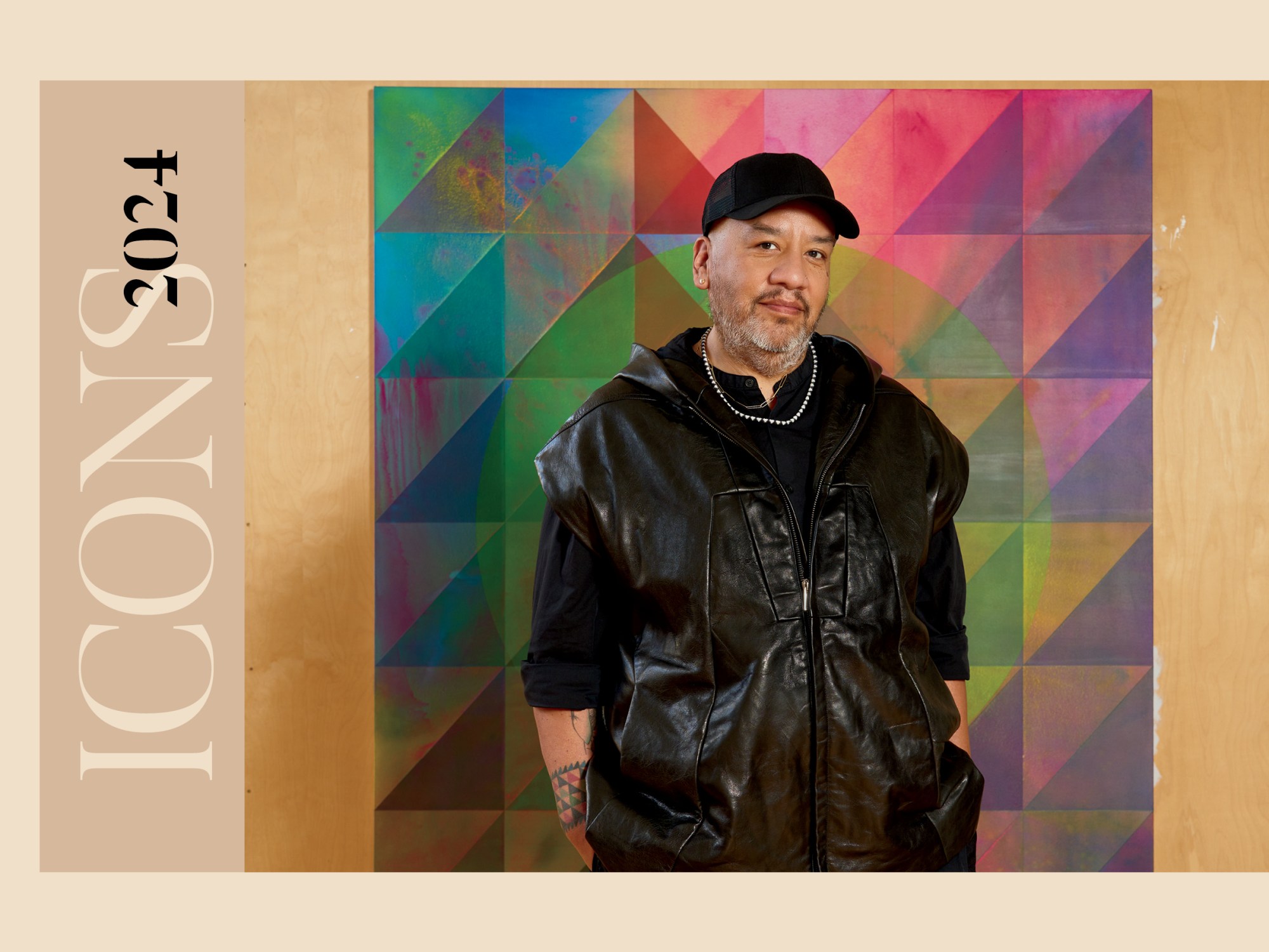 A man in a black vest over a black shirt in a black hat standing in front of a multi-colored abstract painting in the works.