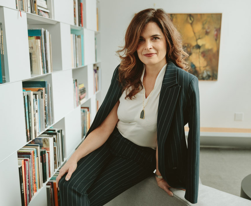 A white woman seated on a low bench. She wears pinstriped pants and a jacket to match. She sits with her legs crossed near a bookshelf.