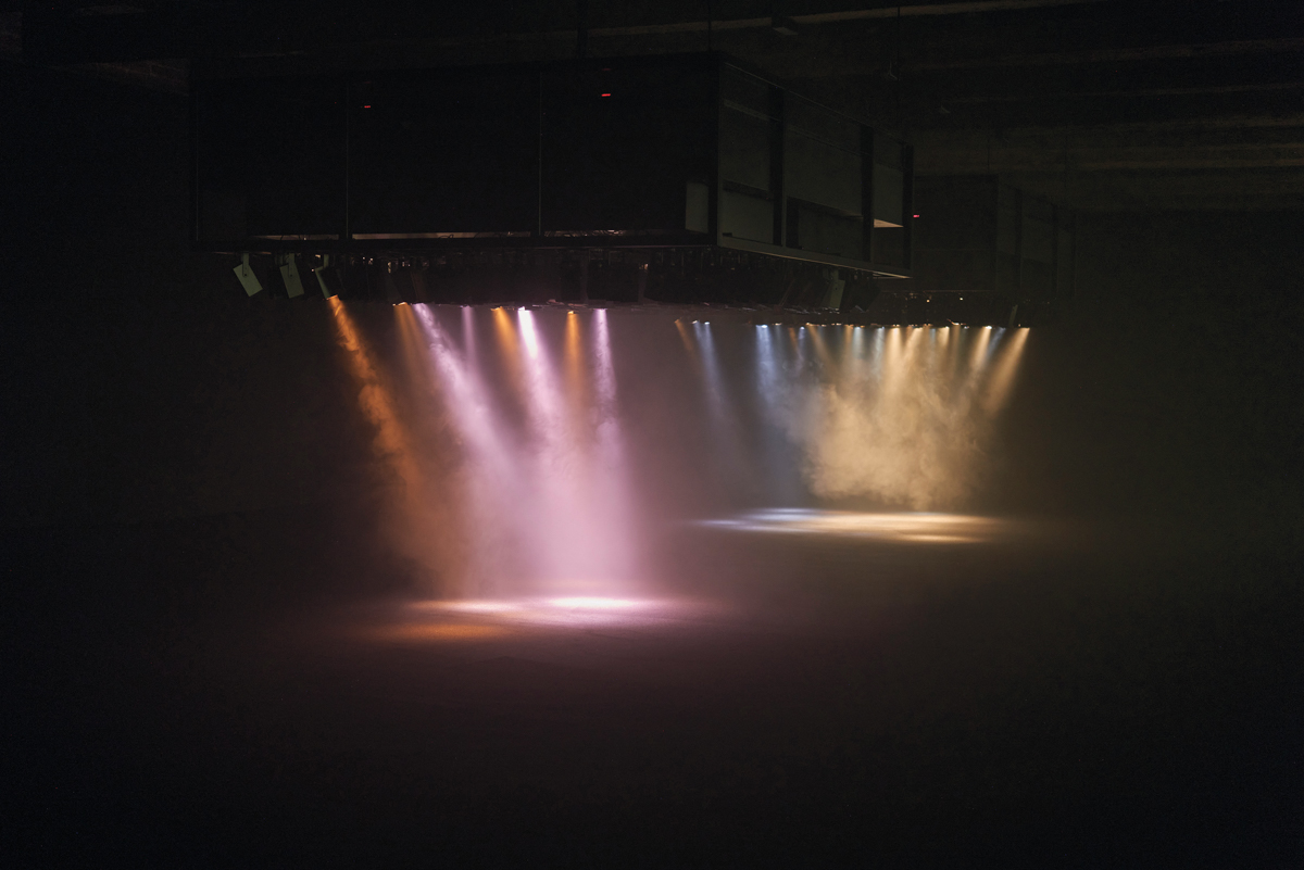 A large darkened room with pink, orange, and yellow beams of light pouring down through haze from a fog machine.