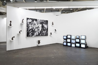 View of an art fair booth showing a black-and-white photograph on the wall surrounded by sculptures on small shelves. At right is a stack of TVs.