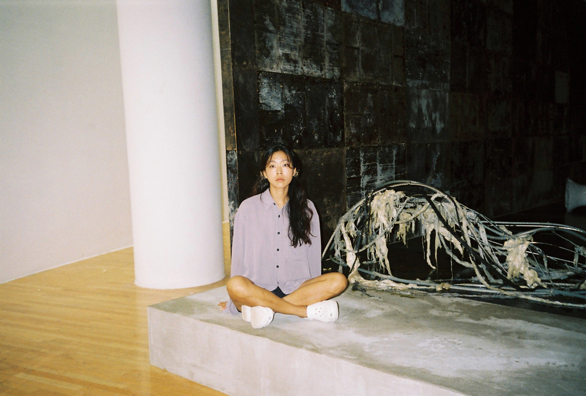 Portrait of Mire Lee, an Asian woman, sitting cross-legged on a concrete platform next of an installation work by her.