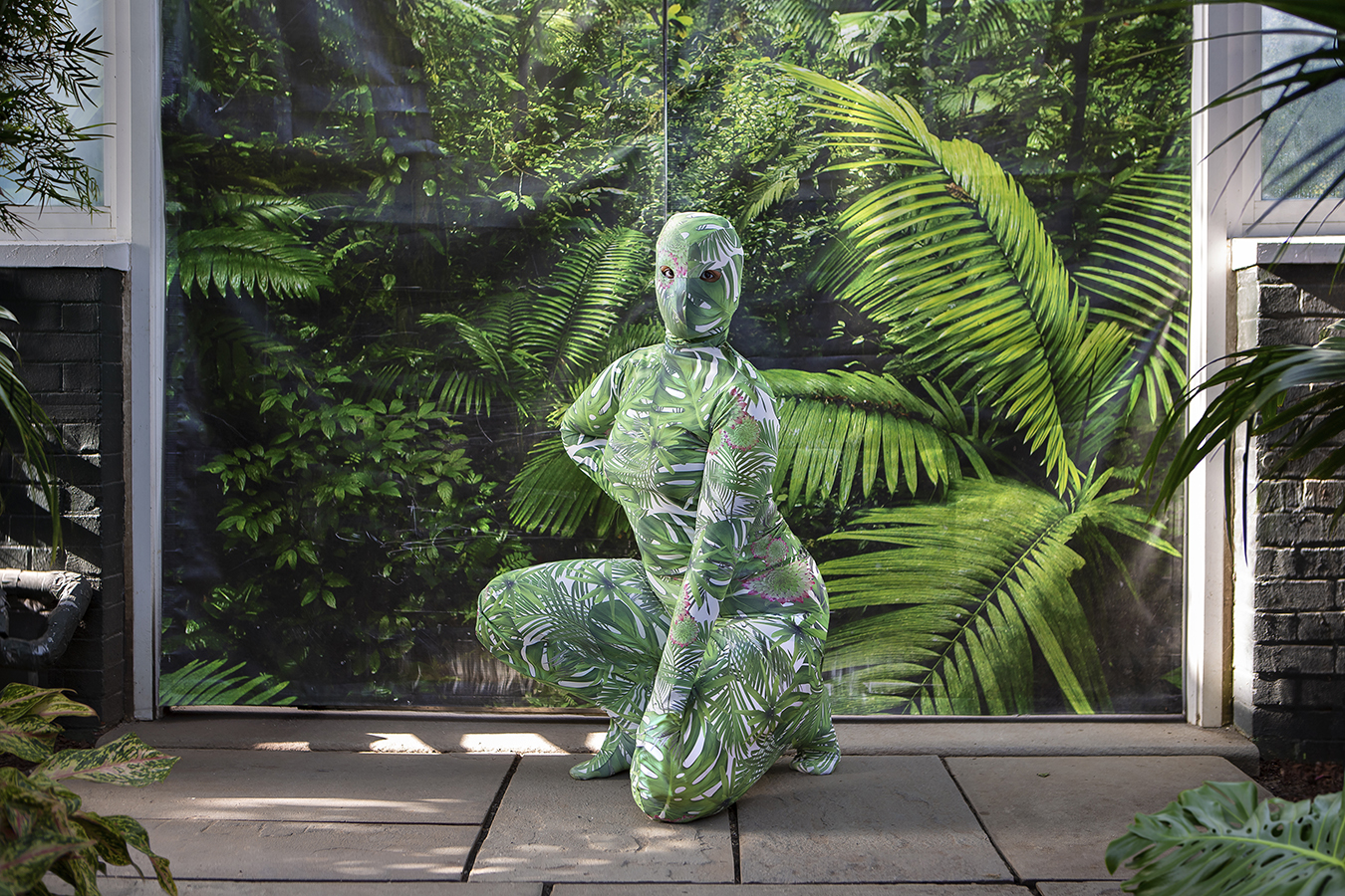 A person wearing a full coverage bodysuit with a tropical print crouches in front of a tropical backdrop.