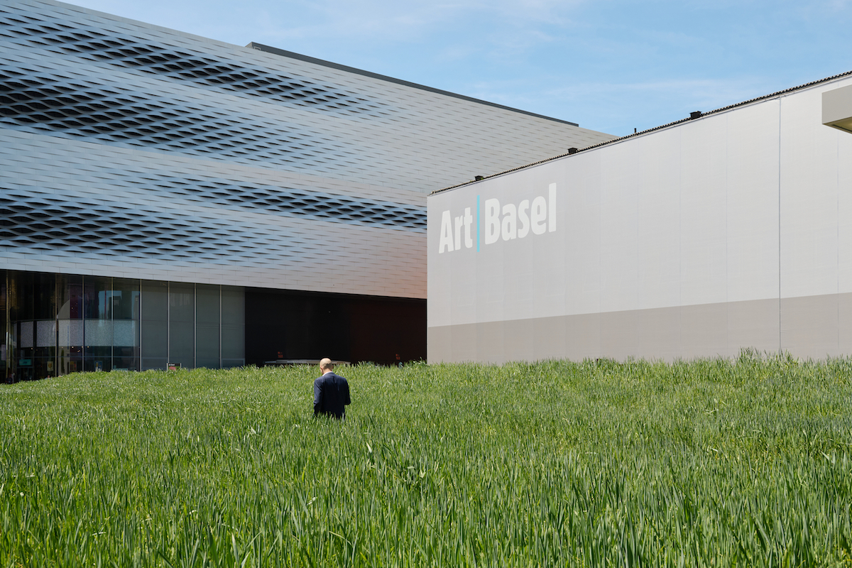 A man standing in a field of wheat before a blocky building with the words 'Art Basel' on its facade.