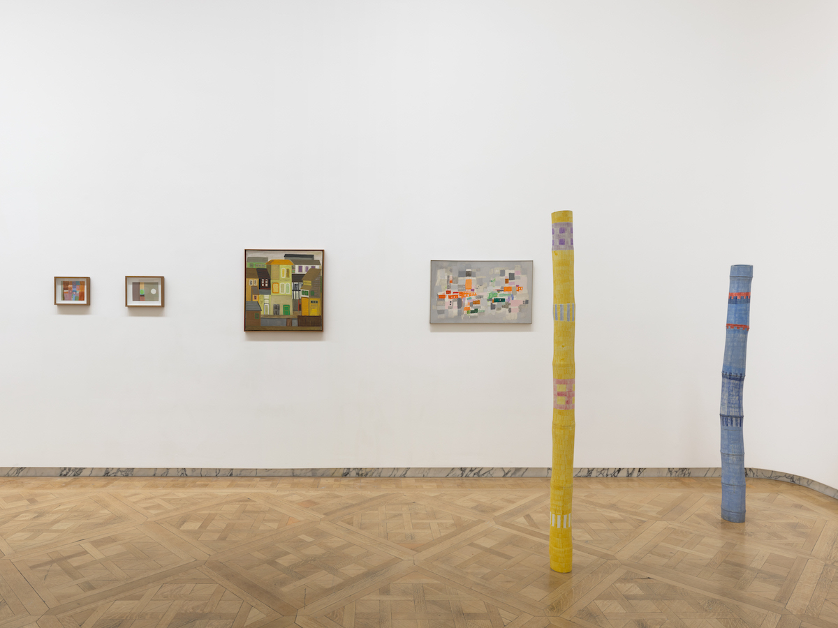 Two-pole like sculptures painted with abstract forms set beside four paintings on a wall.